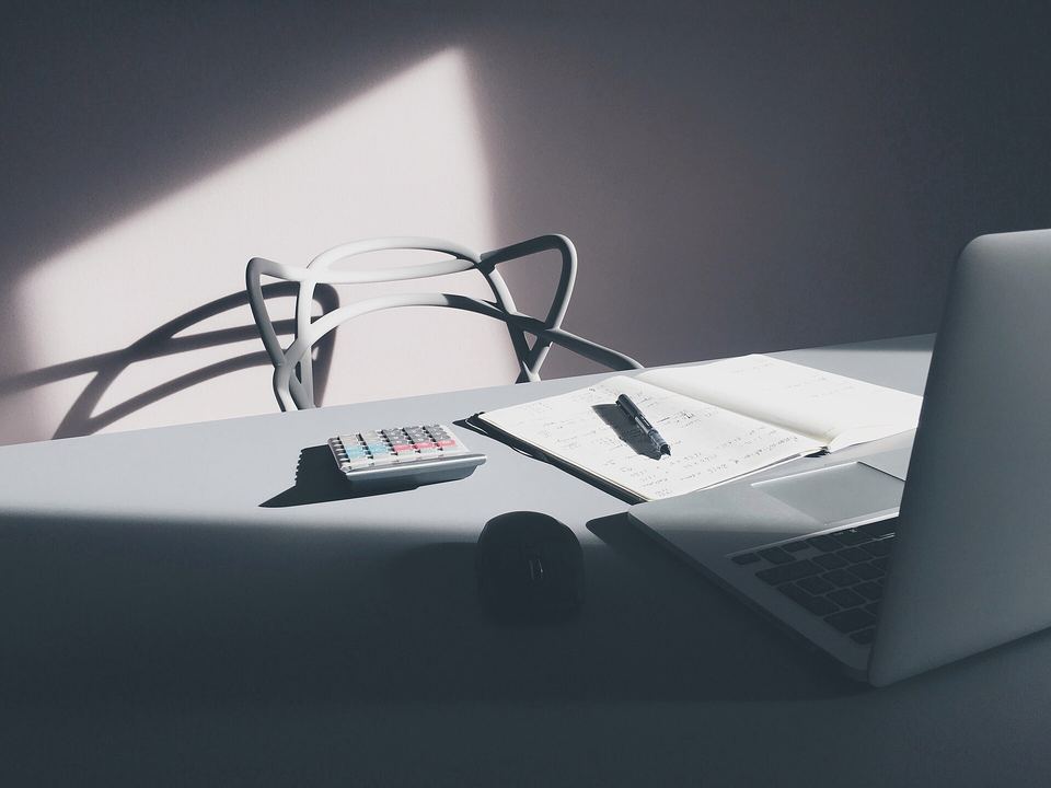 Sunlit desk with a calculator, an open notebook with a pen, a mouse and a laptop
