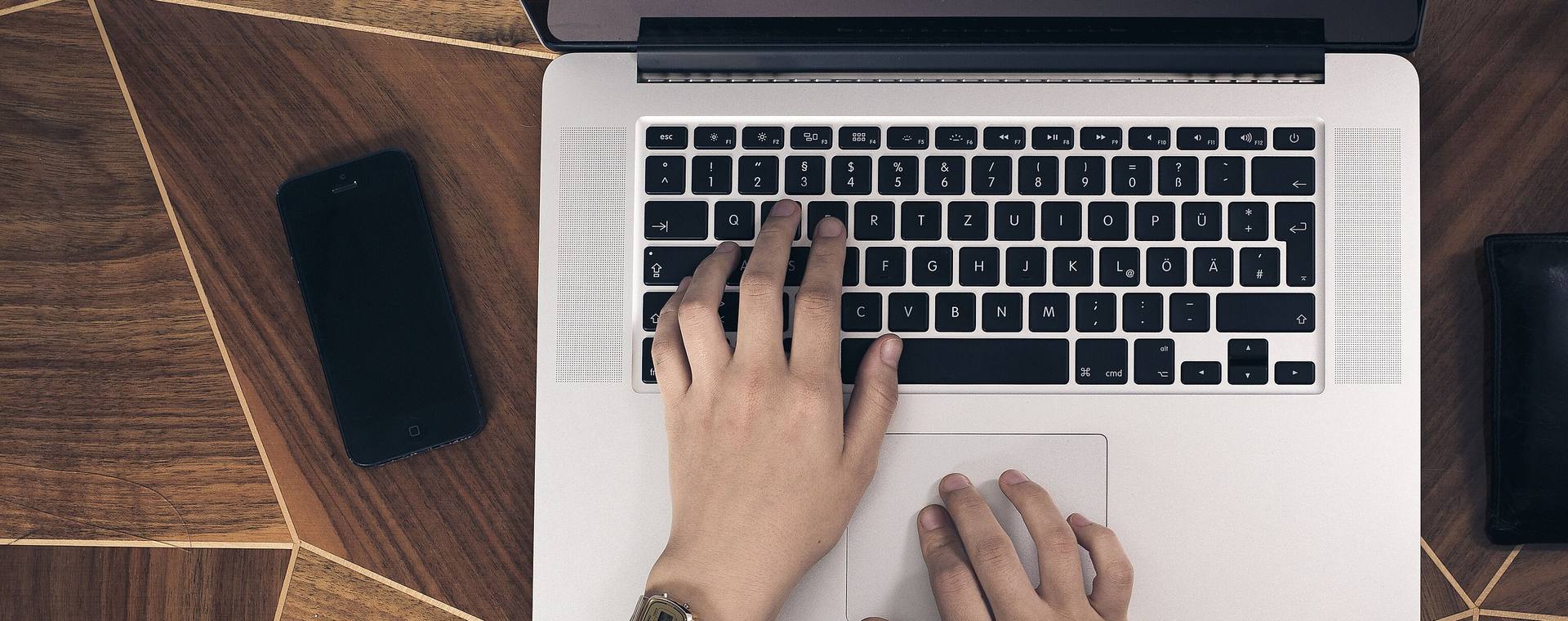 Top view of hands typing on a laptop on a wooden desk next to a smartphone and a leather wallet