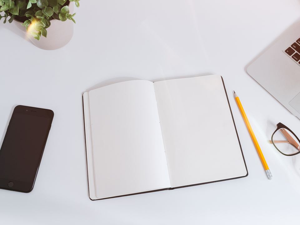 Bright white desk with an open blank notebook, a yellow pencil, glasses, a smartphone and a laptop
